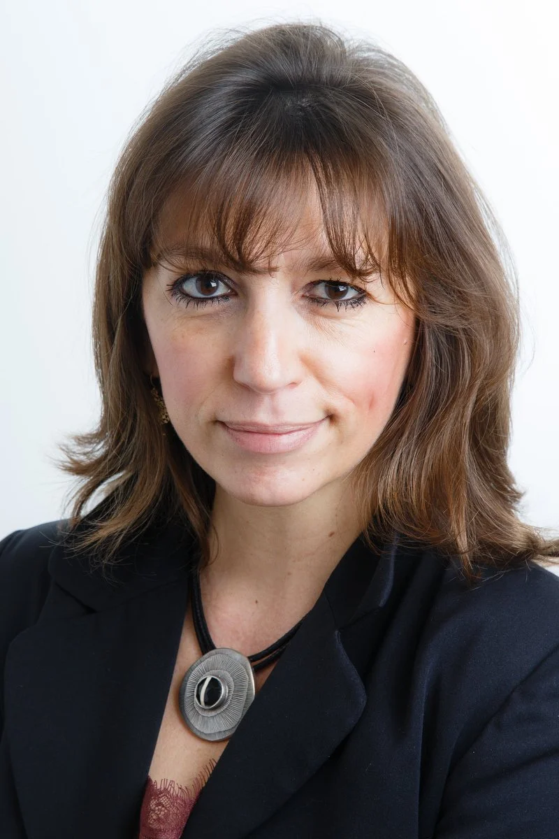 Femme aux cheveux et à la frange bruns, portant un blazer noir et un collier, posant pour un portrait professionnel.