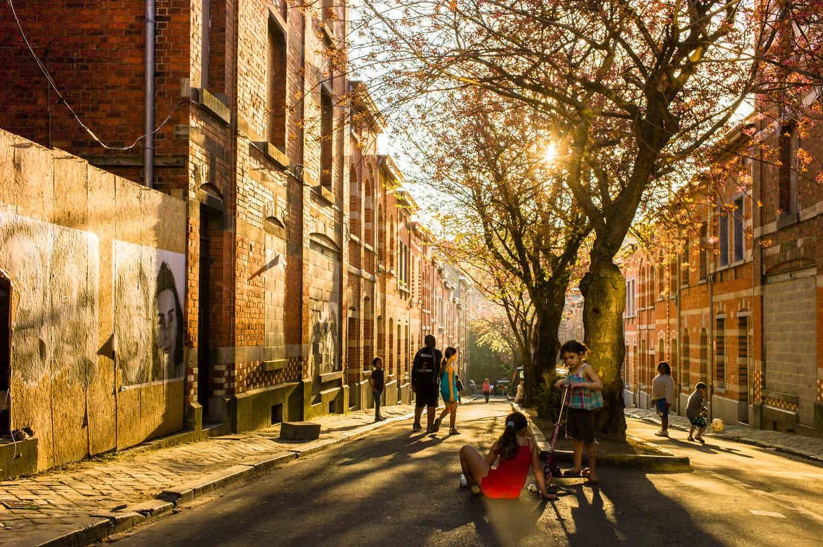 People walking and playing on a sunlit cobblestone street lined with brick buildings and blooming trees.