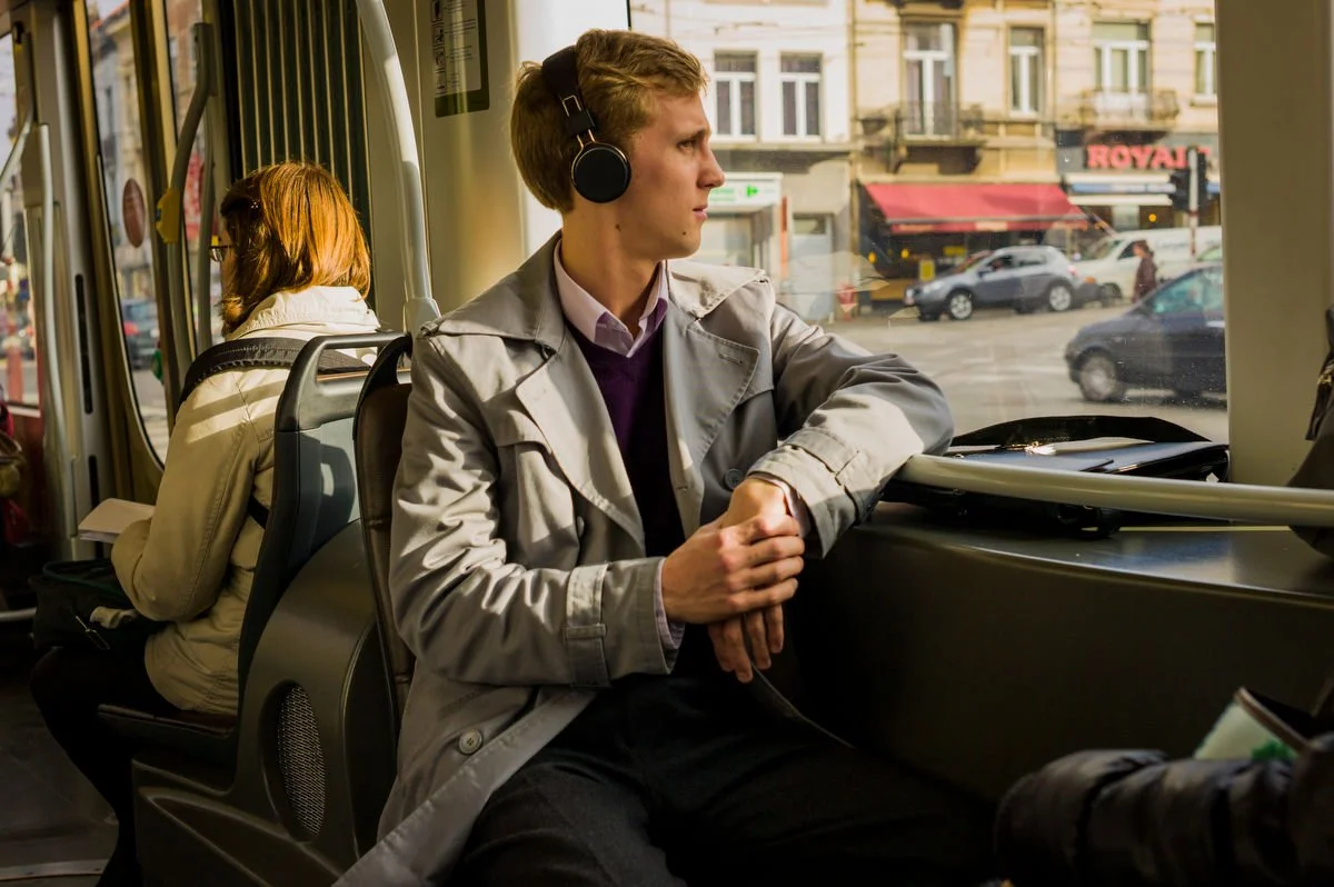 Man wearing headphones sitting on a tram or bus seat, looking out the window, with a briefcase next to him. Another passenger is in the background reading.