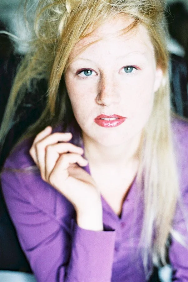 Femme blonde aux yeux bleus vifs et à la chemise violette, la main près du visage, regardant vers la caméra.