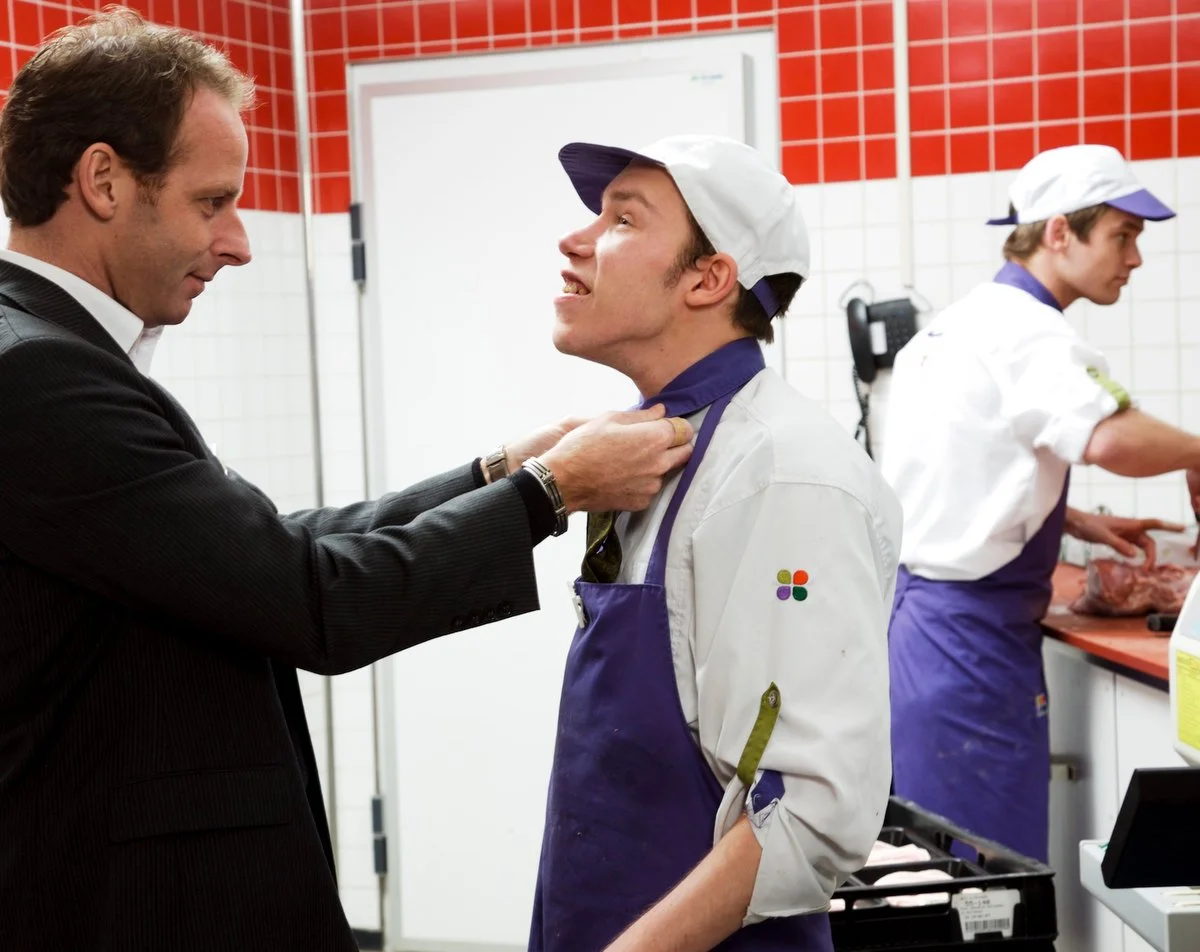 portrait de Deux hommes dans une cuisine. L'un porte un costume sombre et s'agrippe au col de l'autre, qui porte une chemise blanche et un tablier violet. Un troisième homme travaille à l'arrière-plan.