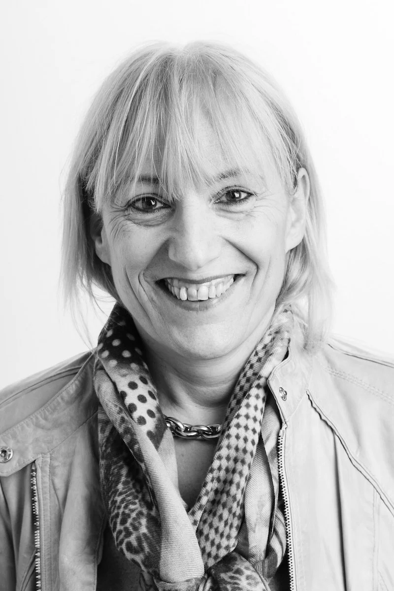 Portrait en noir et blanc d'une personne souriante aux cheveux courts, portant une écharpe à motifs et une veste.