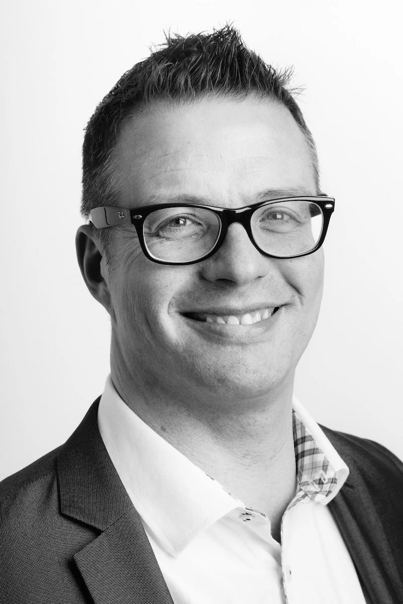 Portrait en noir et blanc d'un homme souriant avec des lunettes, cheveux courts, portant une veste de costume et une chemise.