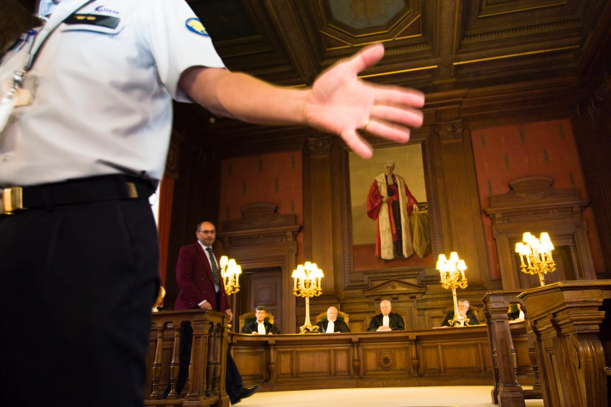 Interior of a courtroom with judges seated at a wooden bench, ornate walls, and a large painting. A person in a uniform is moving in the foreground, obscuring part of the view.