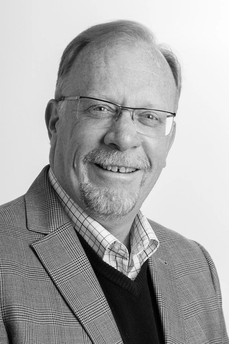 Portrait en noir et blanc d'un homme souriant avec des lunettes, portant une veste de costume à carreaux et une chemise à col.