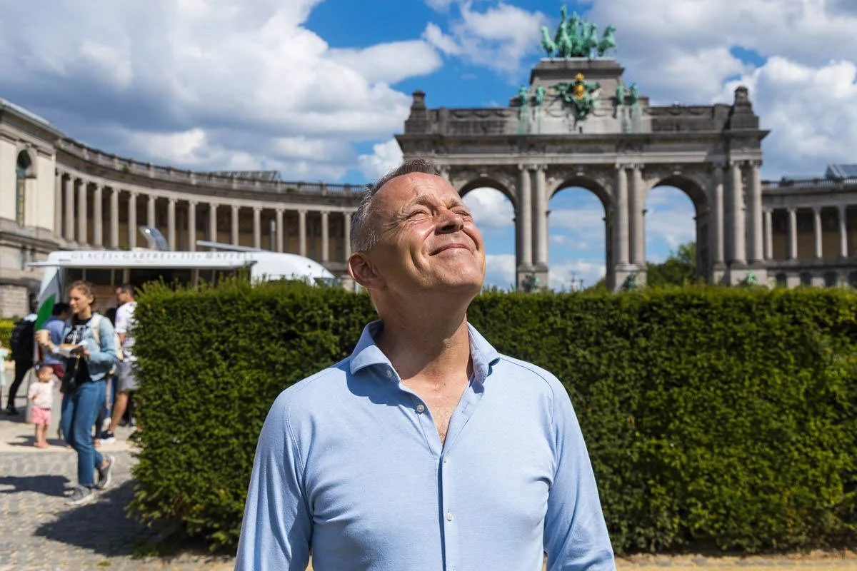Homme en chemise bleu clair debout en plein air devant l'Arc de Triomphe, Bruxelles, sous un ciel bleu avec des nuages.