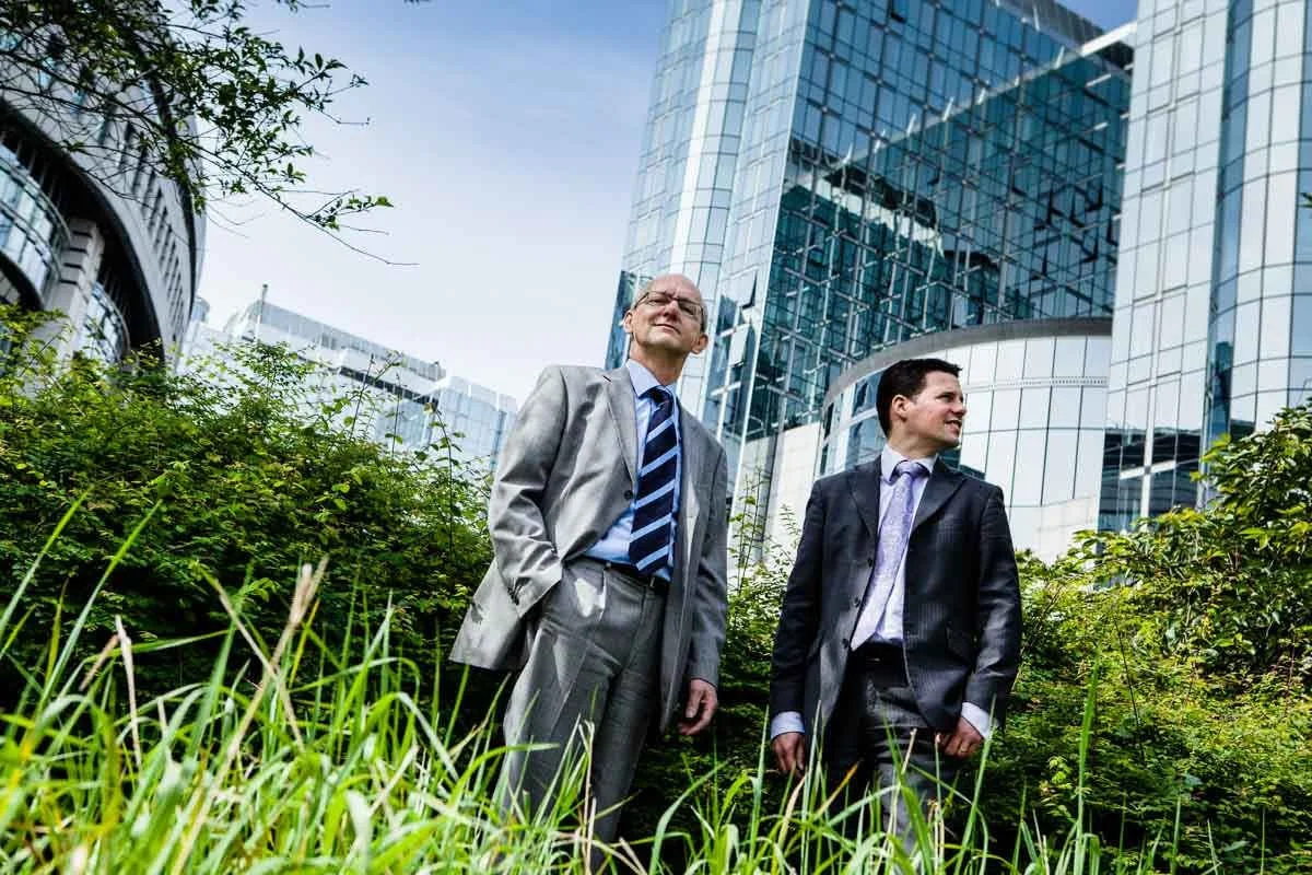 Corporate portrait of two men between the buildings of the European Parliament standing in a grassy part.