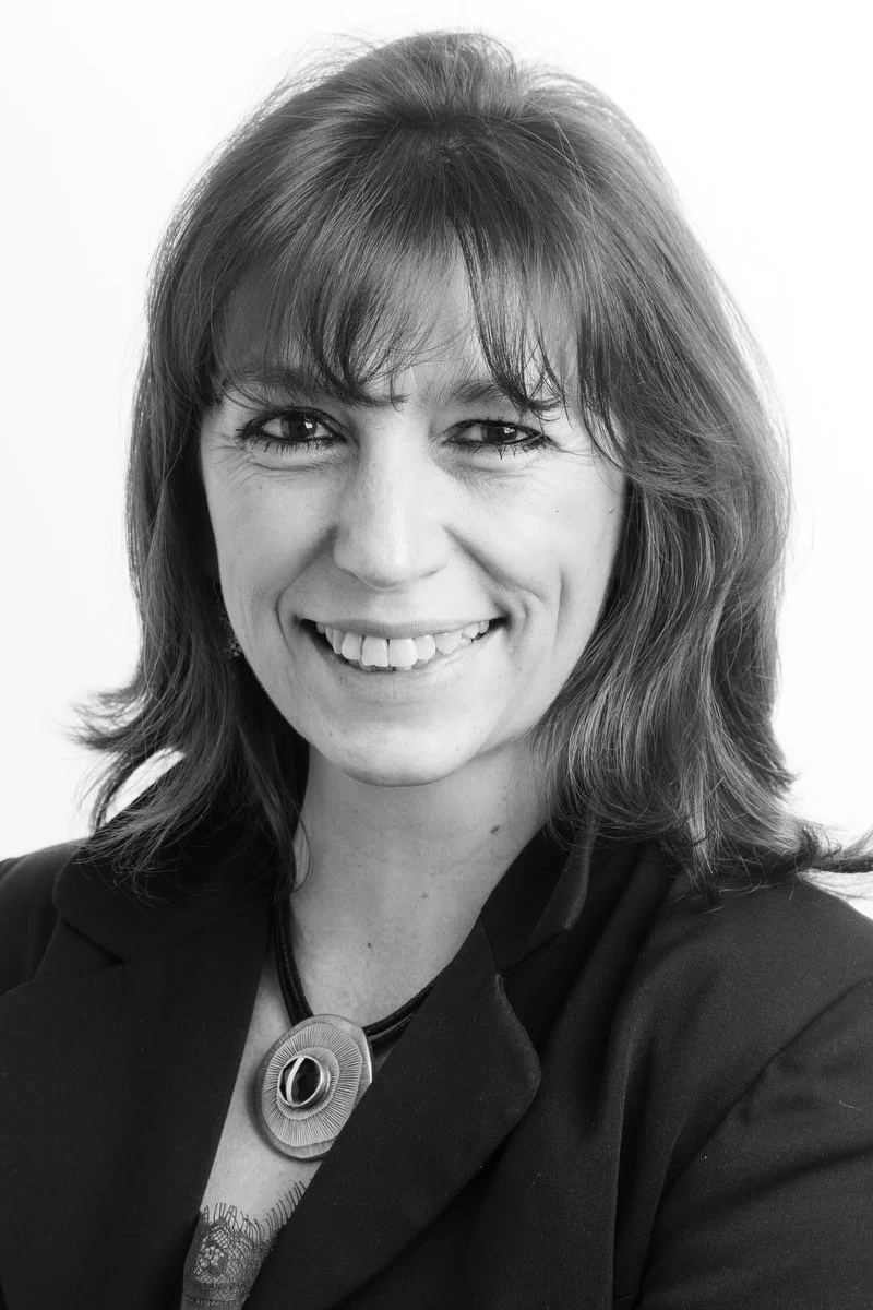 Portrait en noir et blanc d'une femme souriante aux cheveux mi-longs, portant un blazer sombre et un collier.
