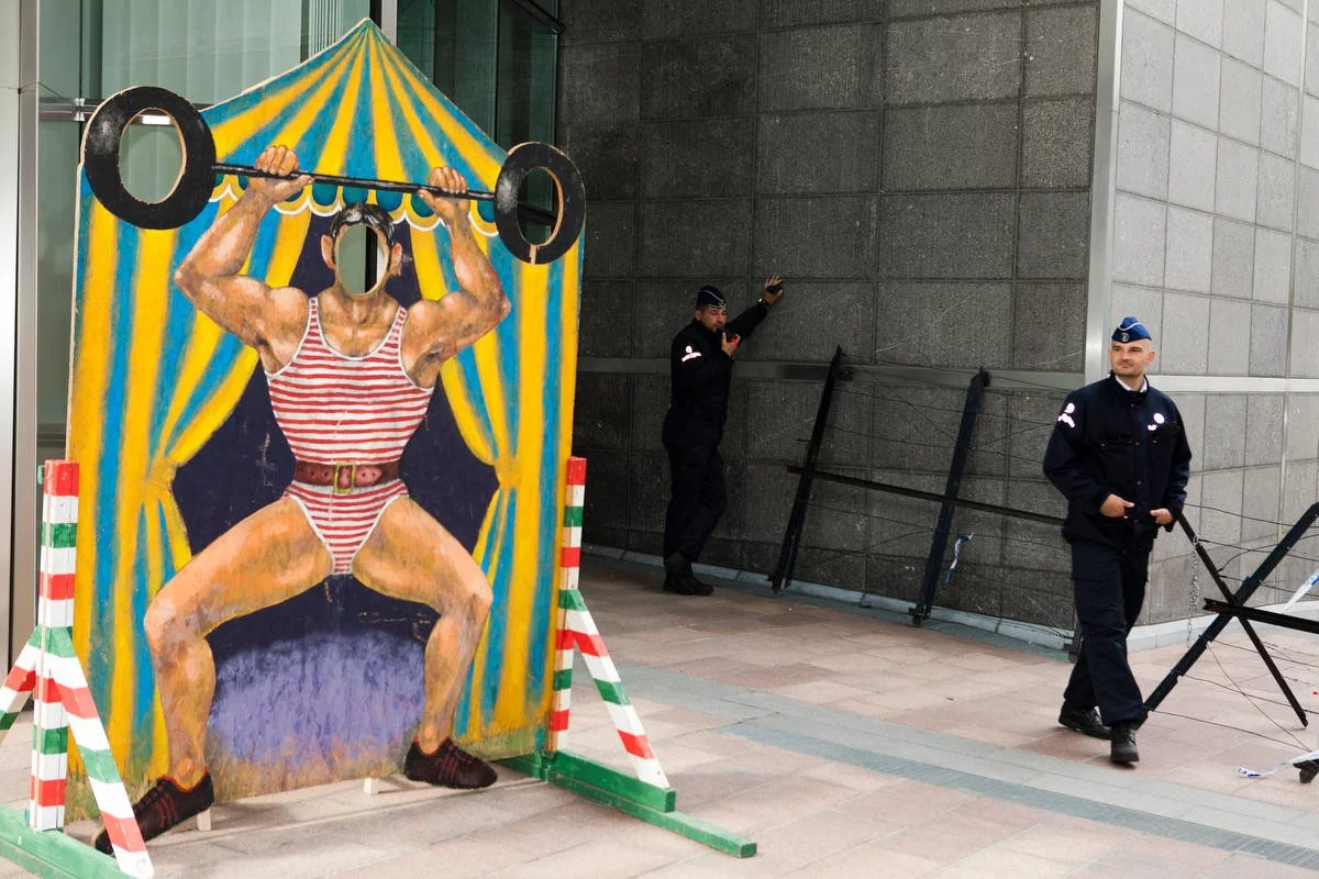 Cutout of a strongman lifting weights with a hole for a face in front of a building, two uniformed officers nearby.