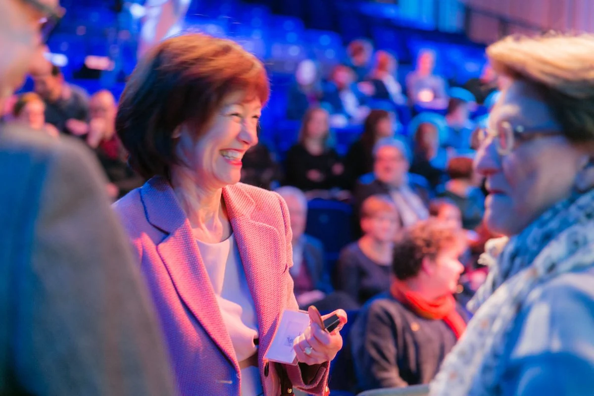 A woman in a pink blazer smiles and chats at an indoor event, captured candidly by a conference photographer in front of a blurred crowd.