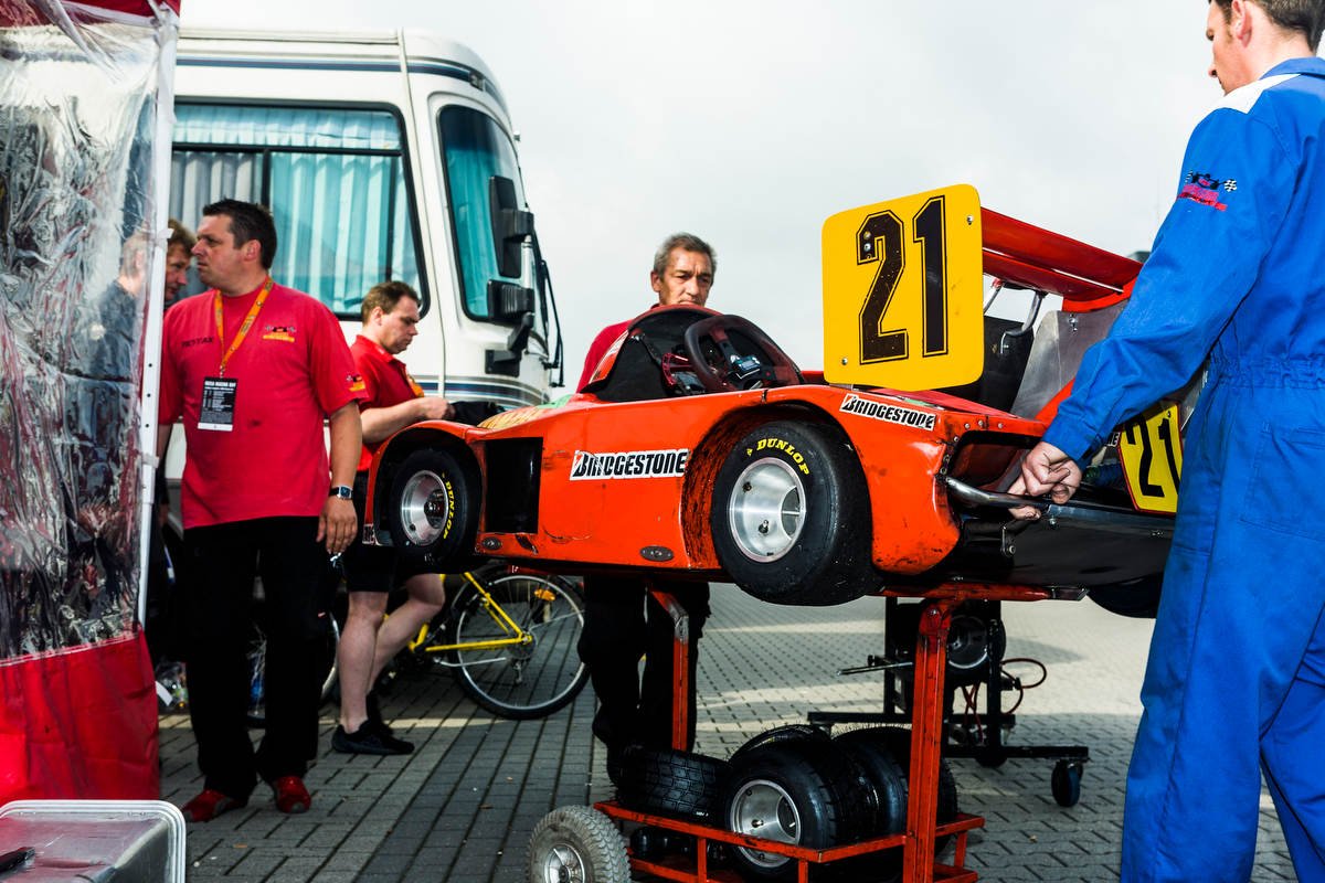 People handling a red mini race car with number 21 in a pit area, near a truck and tents.