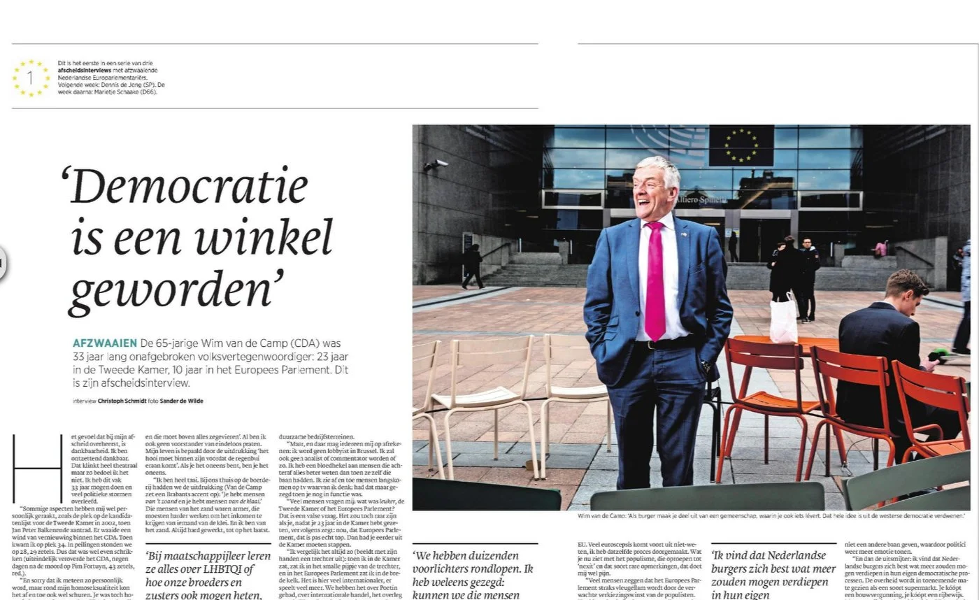 Page de journal présentant un article avec le titre "Democratie is een winkel geworden". Un homme en costume bleu et cravate rouge sourit près de chaises à l'extérieur d'un bâtiment moderne avec un drapeau européen. Un texte en néerlandais entoure l'image.