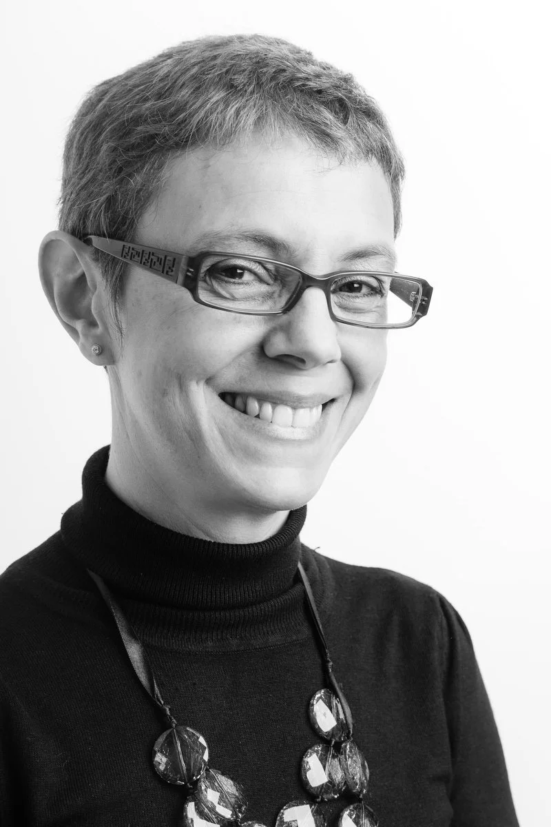 Portrait en noir et blanc d'une personne souriante aux cheveux courts et aux lunettes, portant un col roulé et un collier.