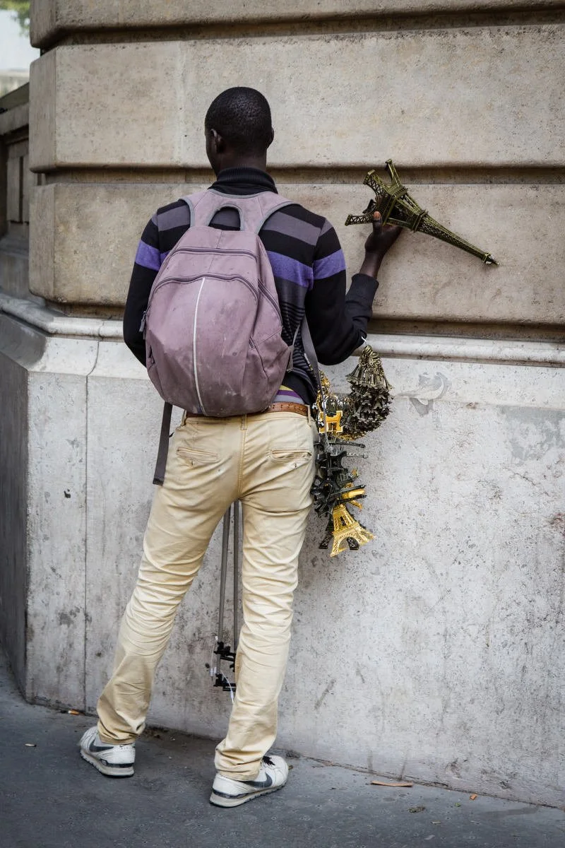 A person with a purple backpack selling miniature Eiffel Tower souvenirs against a stone wall.