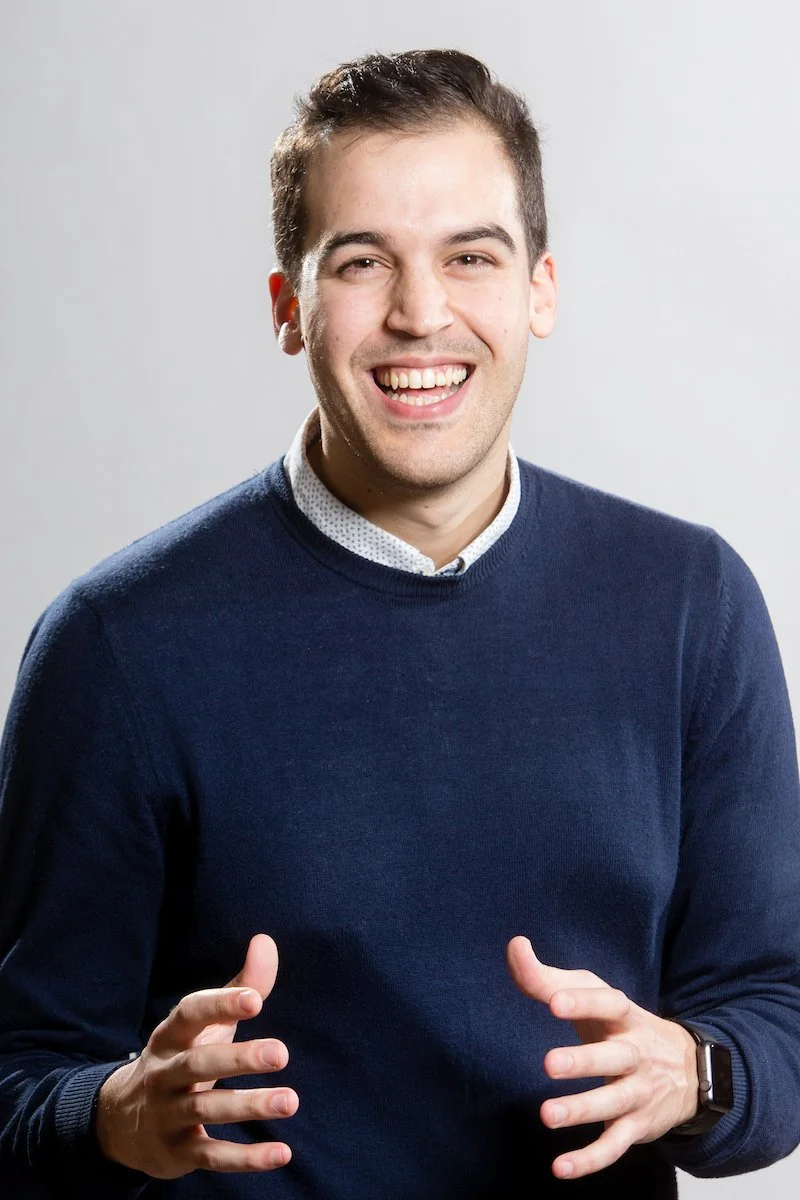 Personne souriante vêtue d'un pull bleu marine, les mains en position de geste.
