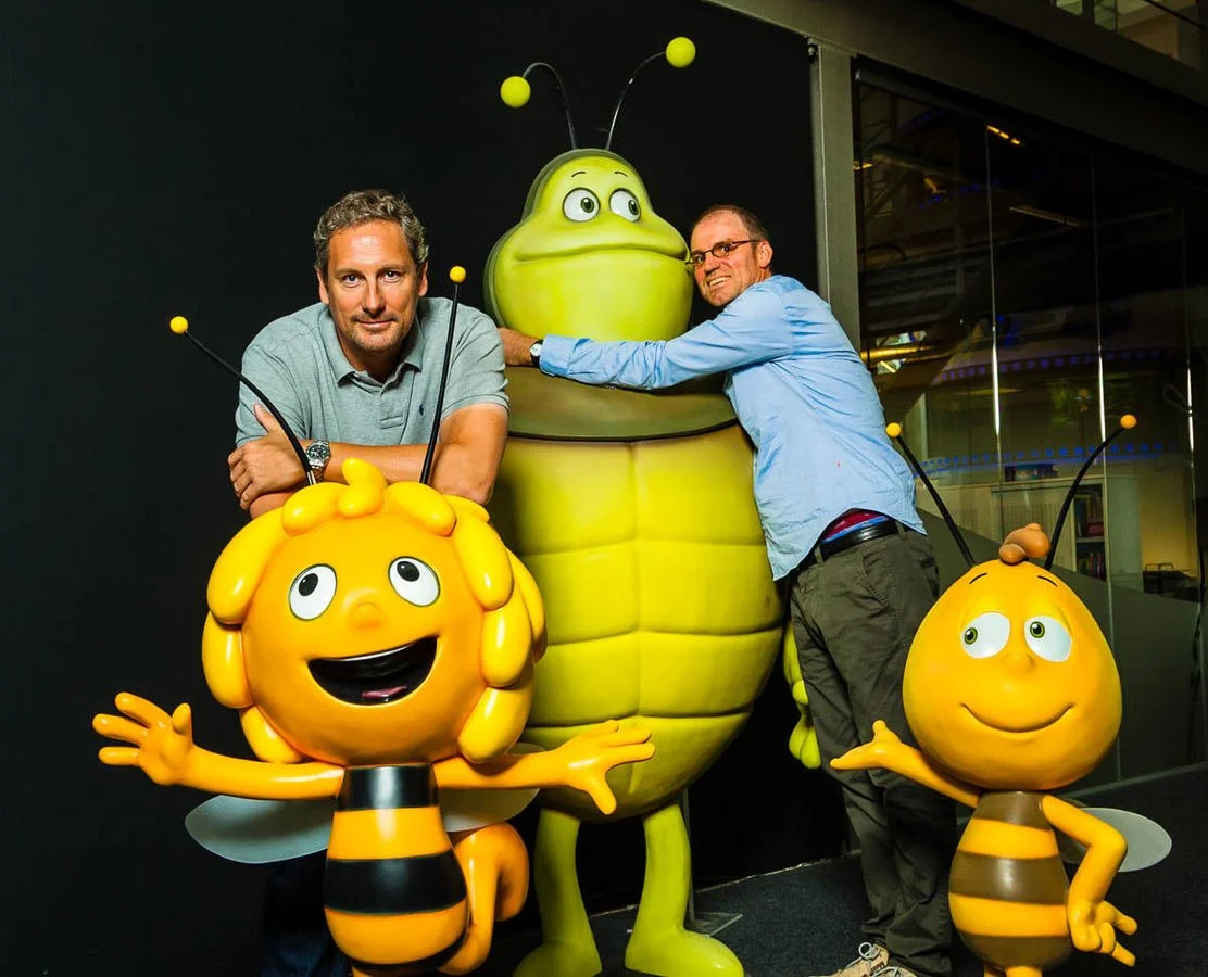 Two men posing with large bee-themed cartoon character statues, including a smiling yellow bee with striped body.