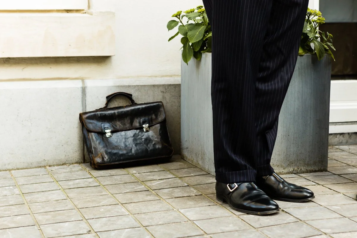 Une personne portant un pantalon à rayures et des chaussures de ville noires se tient à côté d'une jardinière en métal contenant des plantes vertes. Une mallette en cuir usée est posée sur le sol à proximité.