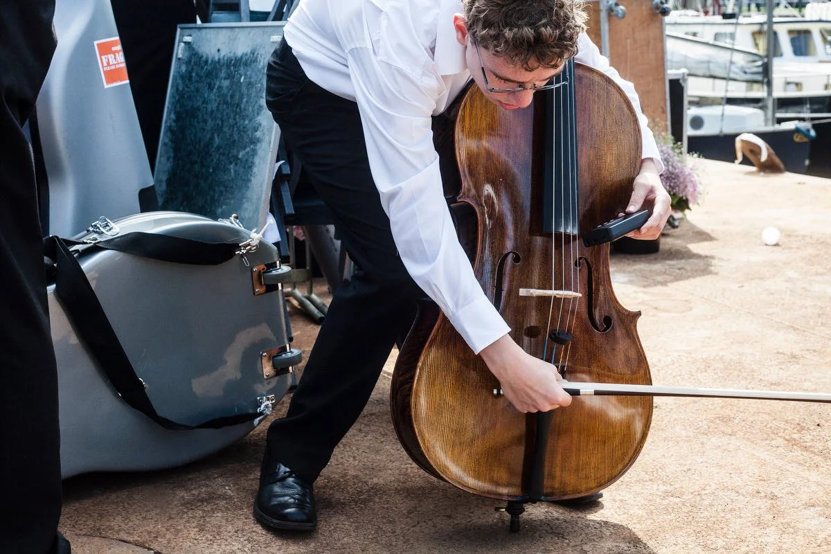A person wearing formal attire tuning a cello outdoors, next to a cello case, with a dock and boats in the background.