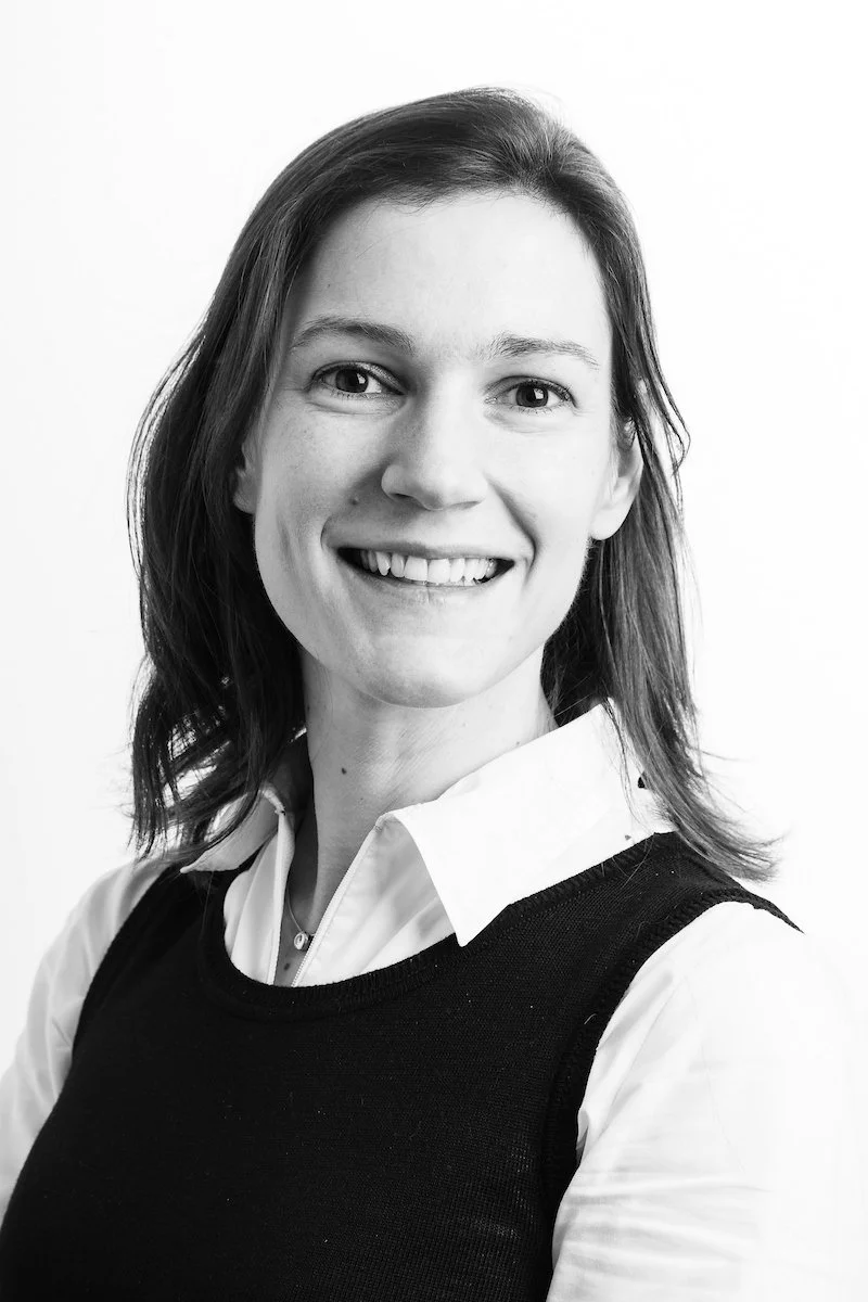 Portrait en noir et blanc d'une femme souriante aux cheveux mi-longs, vêtue d'une chemise blanche et d'un gilet noir.
