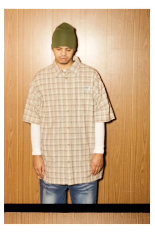 Personne portant un bonnet vert et une chemise à carreaux sur une manche longue blanche, debout contre un mur en bois.