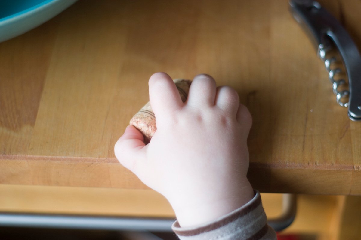 Child's hand holding a wine cork on a wooden table next to a corkscrew.