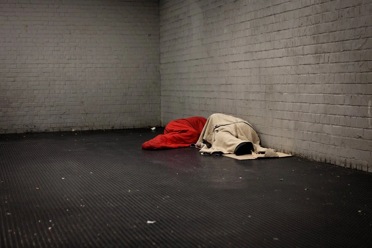 A homeless person sleeping in a corner, covered with a beige blanket and red sleeping bag on a cardboard mat, near a brick wall.