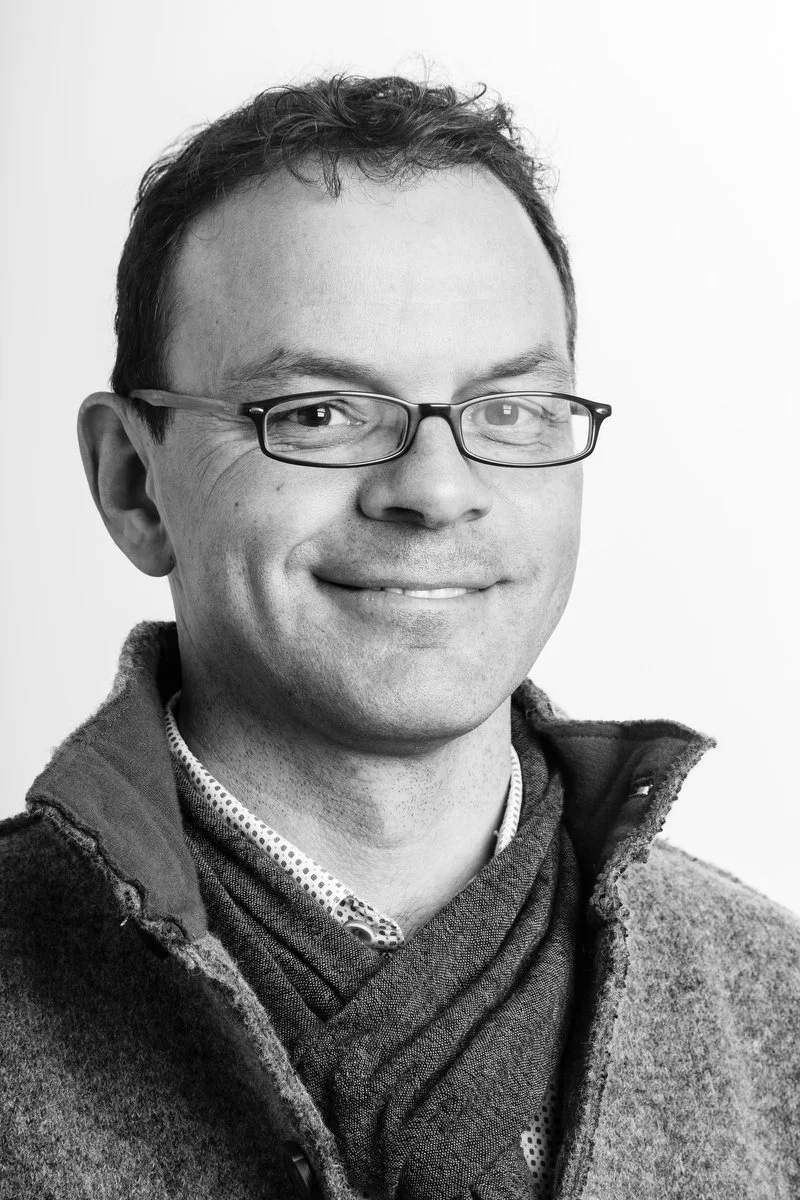 Portrait en noir et blanc d'un homme portant des lunettes, une chemise à col et un pull à col montant, souriant à l'appareil photo.