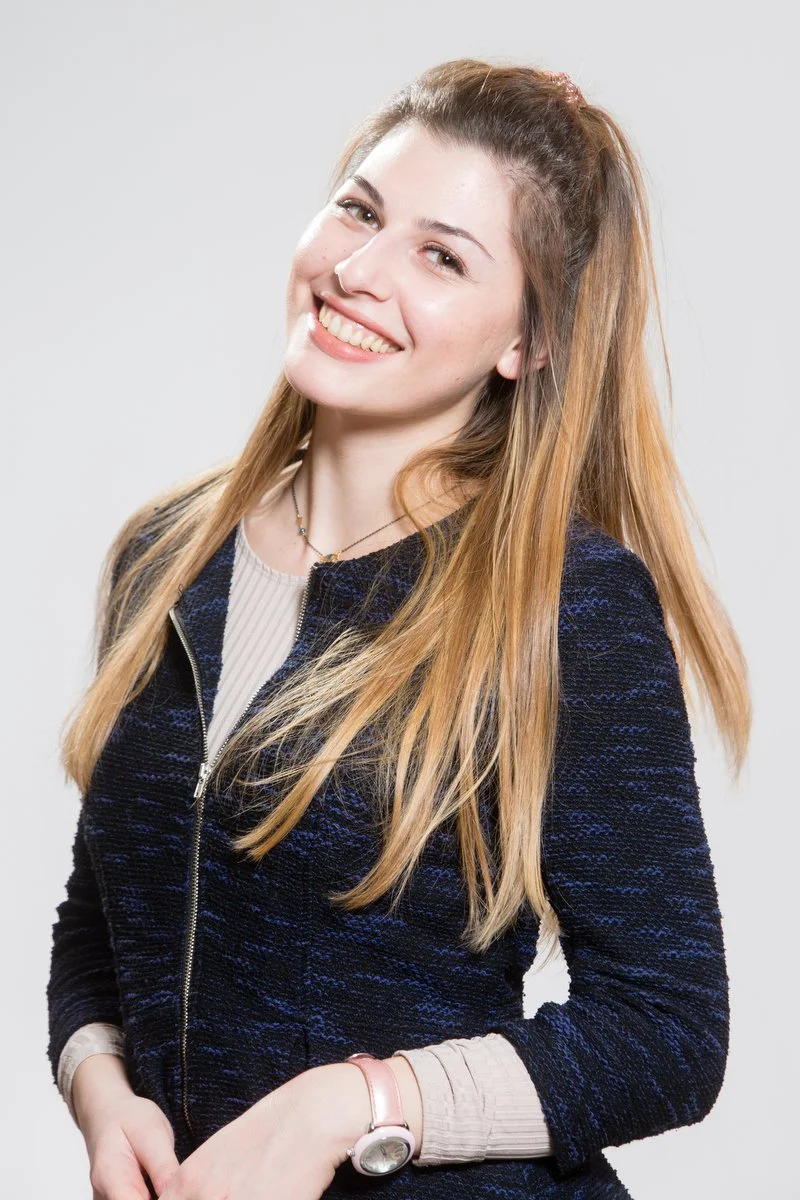 Personne souriante aux cheveux longs, portant une veste sombre, posant sur un fond clair.