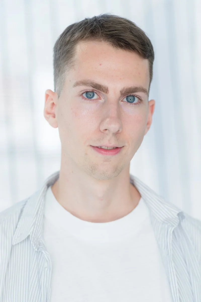 Portrait d'une personne à l'expression neutre, vêtue d'une chemise blanche et d'une surchemise rayée, sur fond clair.