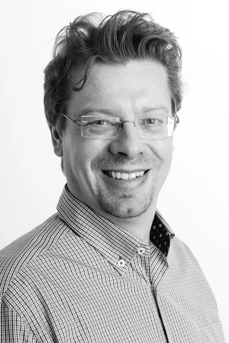Portrait en noir et blanc d'un homme souriant portant des lunettes et une chemise à carreaux.