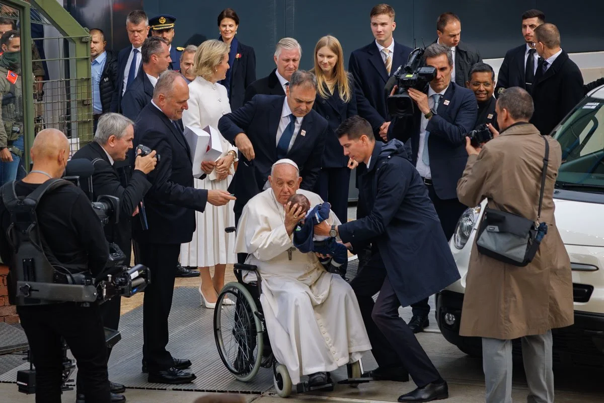 Un homme en robe blanche assis dans un fauteuil roulant, entouré d'un groupe de personnes, dont des photographes et des membres du personnel de sécurité, et tenant un bébé dans ses bras. Des hommes et des femmes vêtus de costumes et de robes observent la scène.