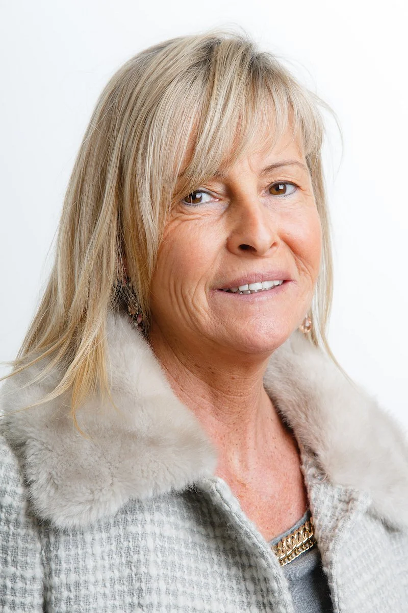Femme aux cheveux blonds vêtue d'un manteau à carreaux clair avec un col en fourrure, souriante.