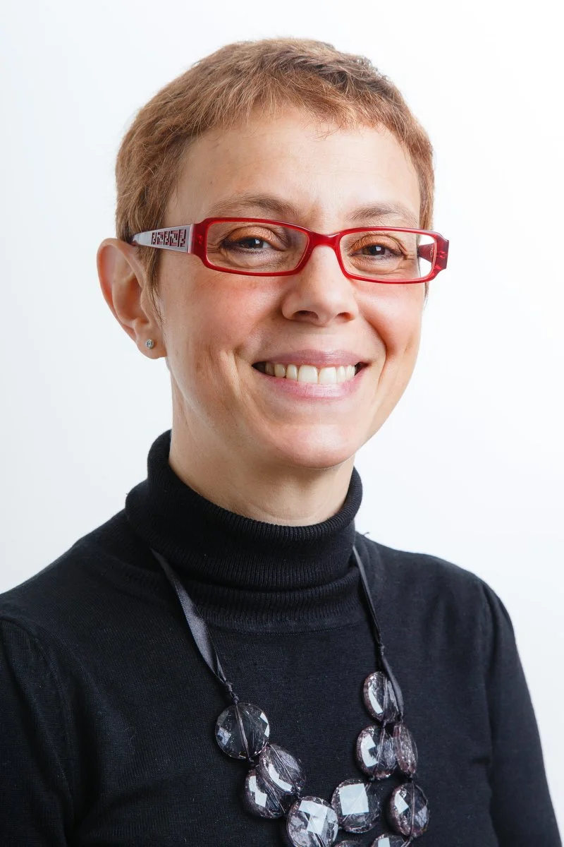 Personne portant des lunettes rouges, un col roulé noir et un collier de perles, souriante.