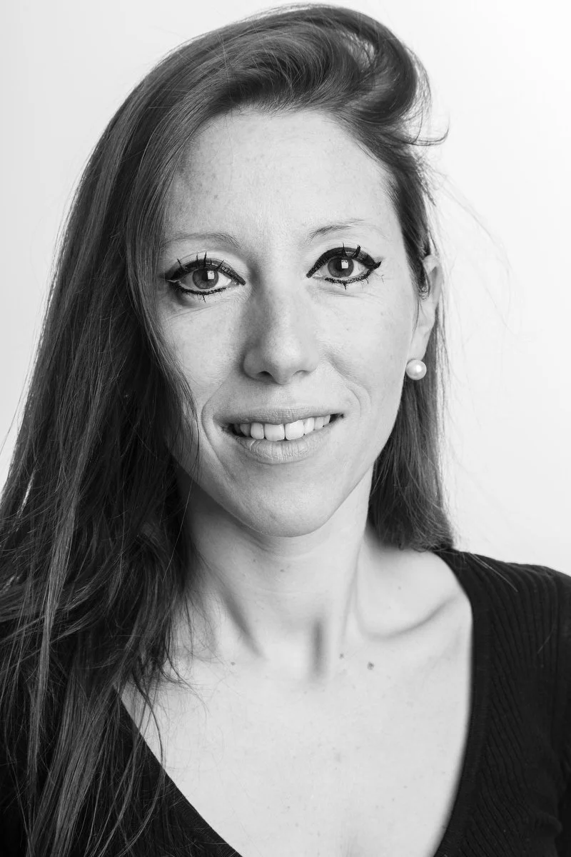 Portrait en noir et blanc d'une femme aux cheveux longs, portant des boucles d'oreilles en perles et un eye-liner foncé, souriant à l'appareil photo.