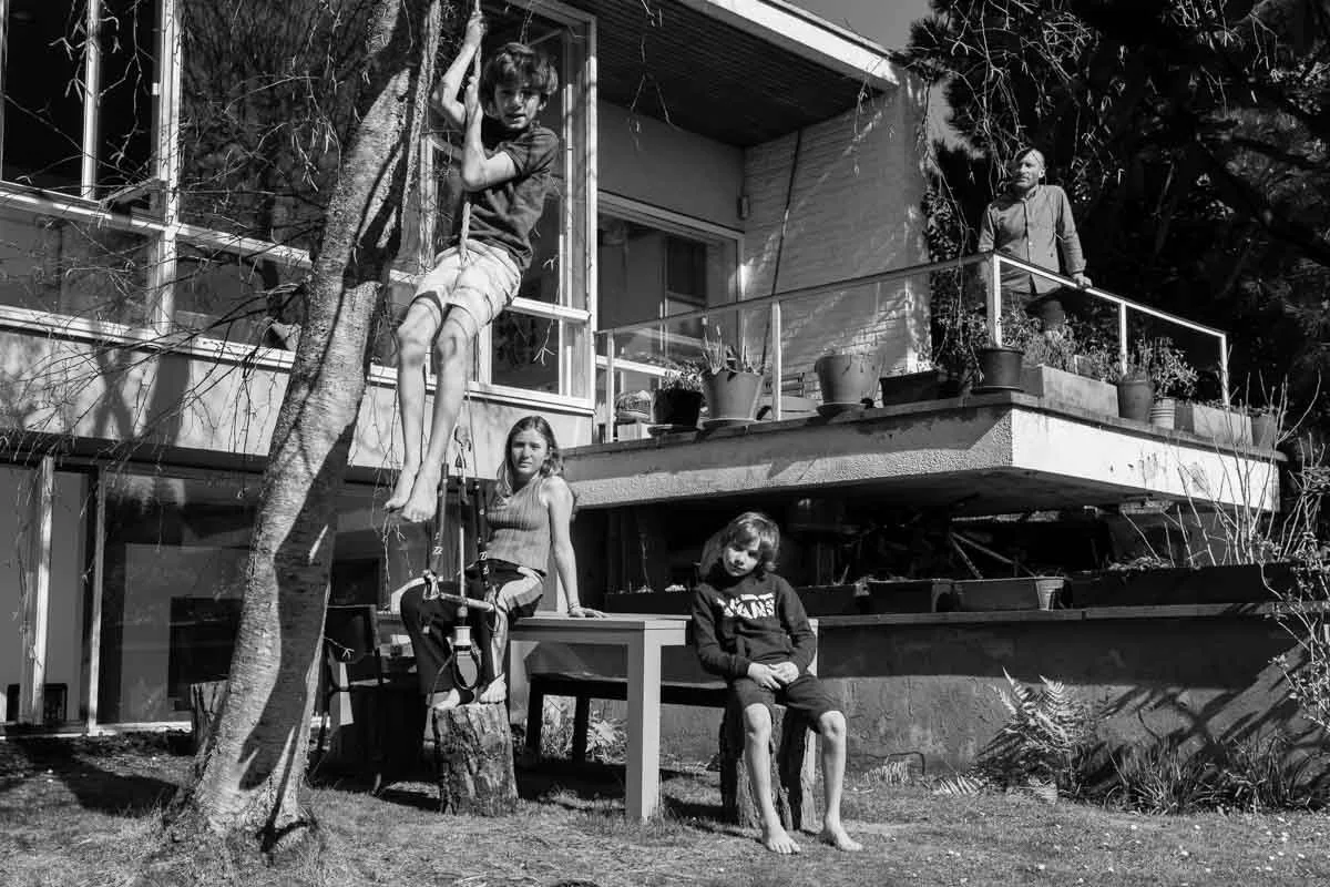 Photo en noir et blanc de trois enfants et d'un adulte devant une maison moderne. Un enfant grimpe à un arbre, un autre est assis sur une table et un troisième est assis sur une souche d'arbre. L'adulte est sur un balcon avec des plantes.