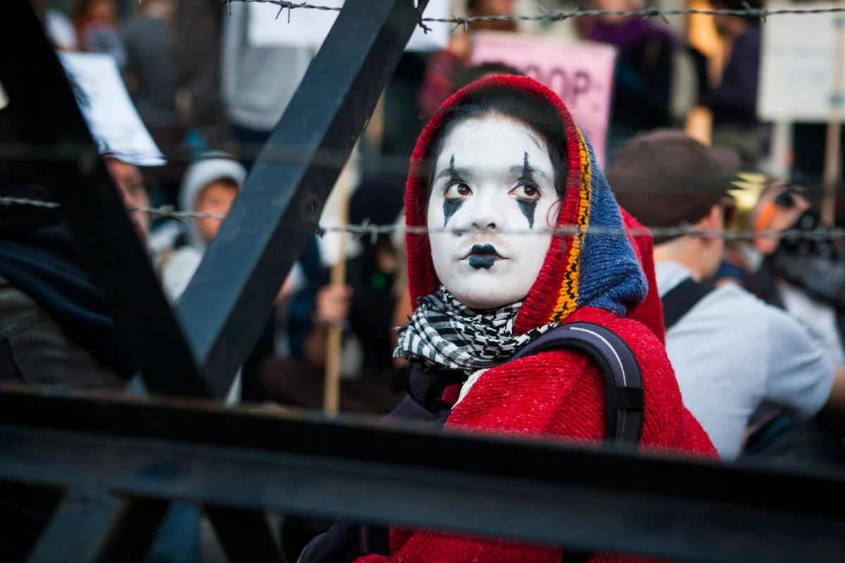 Personne maquillée en clown et vêtue d'une cagoule rouge lors d'une manifestation derrière une clôture de barbelés, avec des personnes tenant des pancartes à l'arrière-plan.