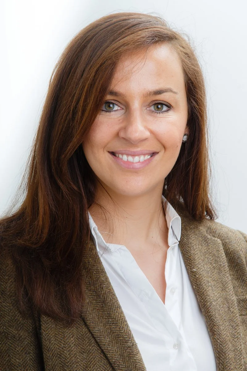 Femme souriante aux longs cheveux bruns portant un blazer brun et une chemise blanche.