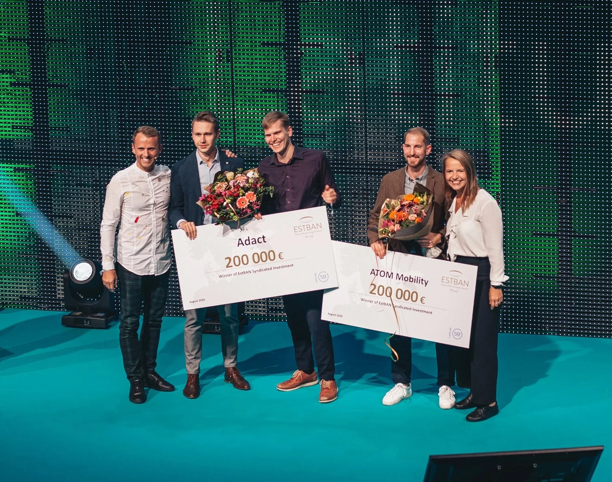 Five people standing on a stage, celebrating and holding large checks and bouquets of flowers, with a colorful LED background and stage lighting.