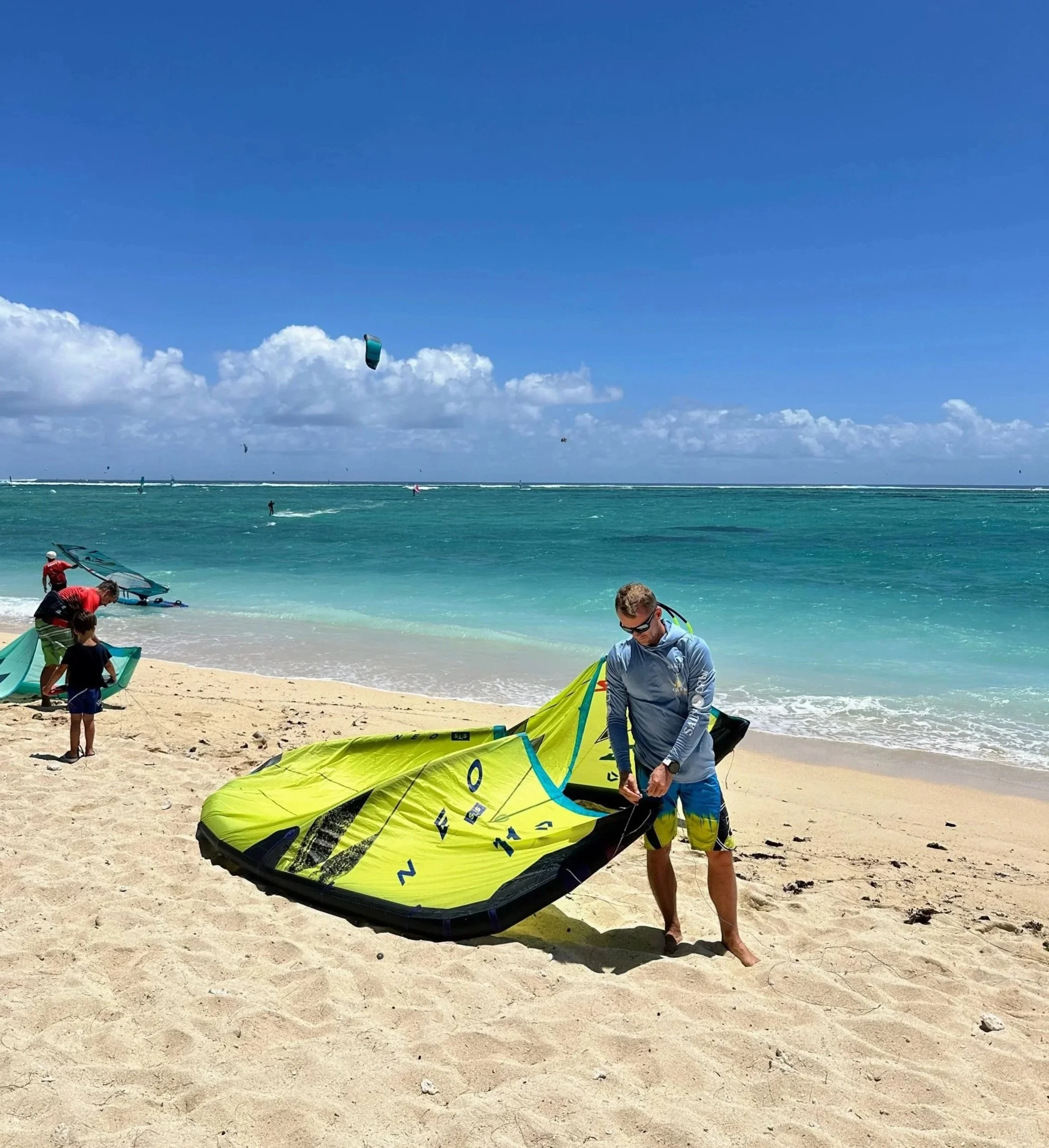 A man prepares a yellow and black kiteboarding kite on a sandy beach with turquoise waves and blue sky in the background. Other kiteboarders and a kite in the air are visible in the distance.