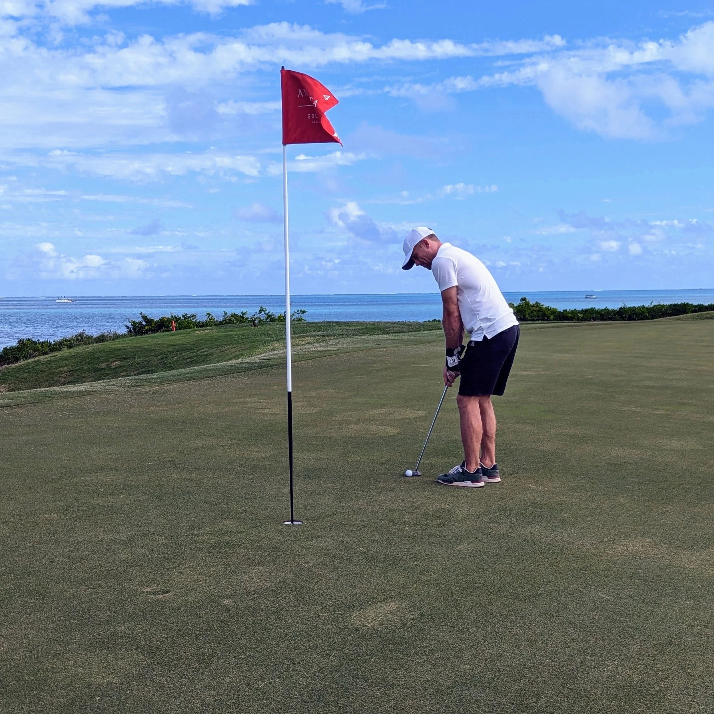 Man preparing to putt on a golf course near the ocean, with a red flag marking the hole, under a partly cloudy sky.