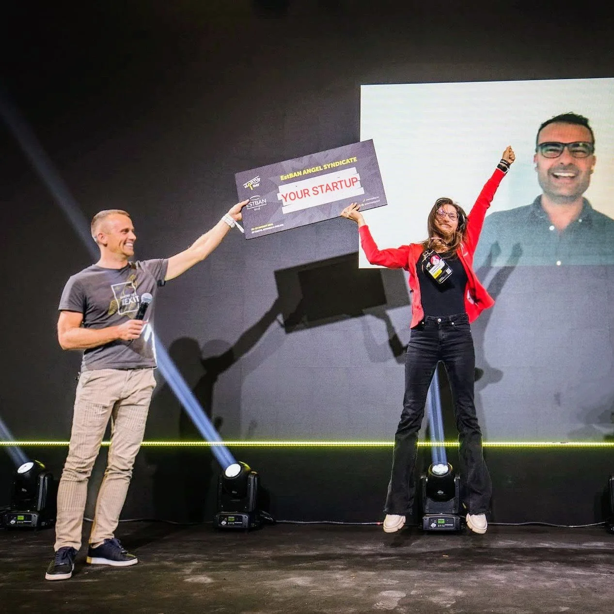 Two people on stage, one holding a microphone and the other jumping with arms raised, holding a large check that reads 'Your Startup'. A large screen behind them displays a man's face.