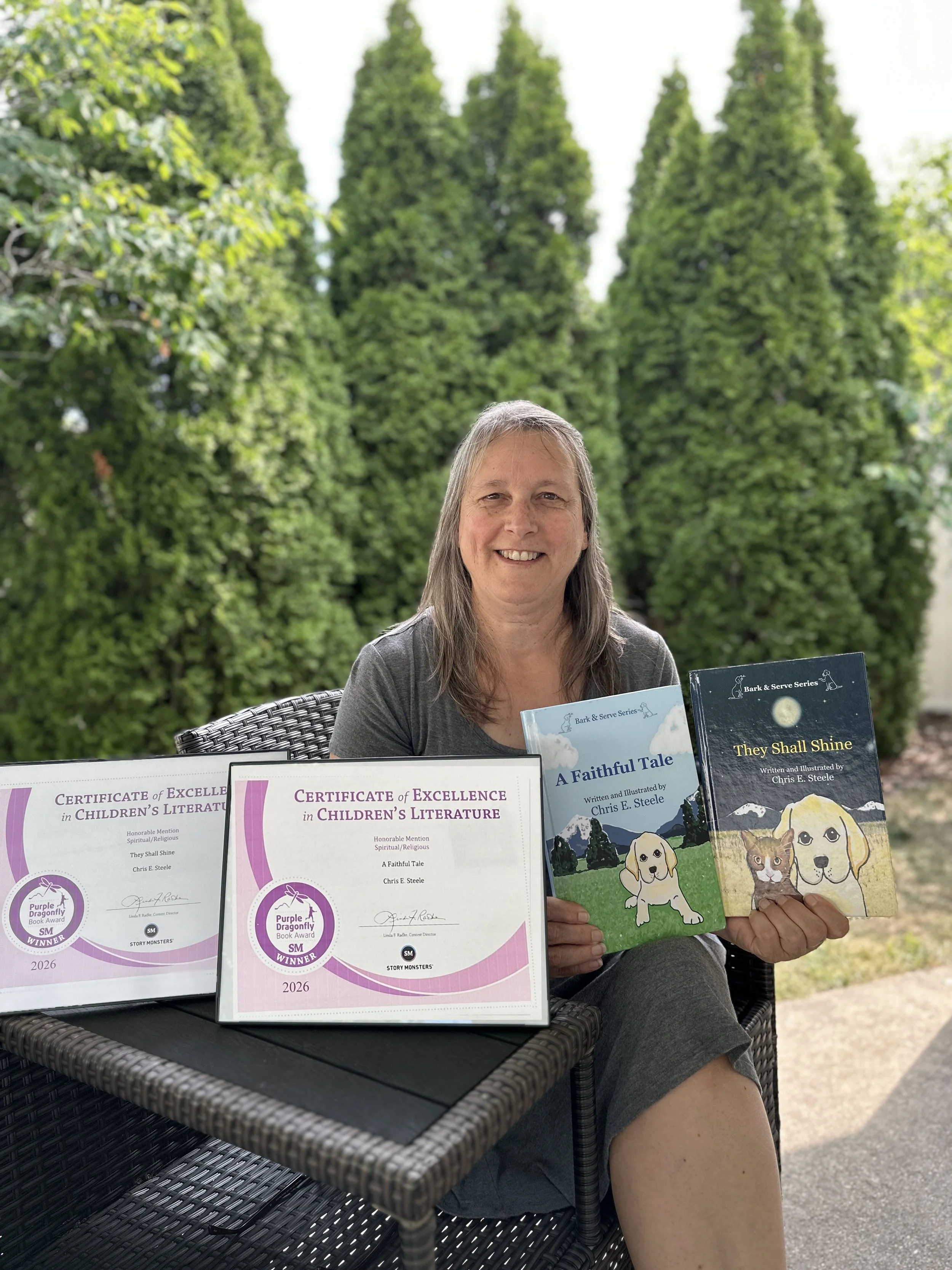 A woman sitting at a table outdoors with a background of green trees, holding books in her hands. On the table are certificates of excellence for children's literature and two books, one titled "A Faithful Tale" and the other "They Shall Shine".