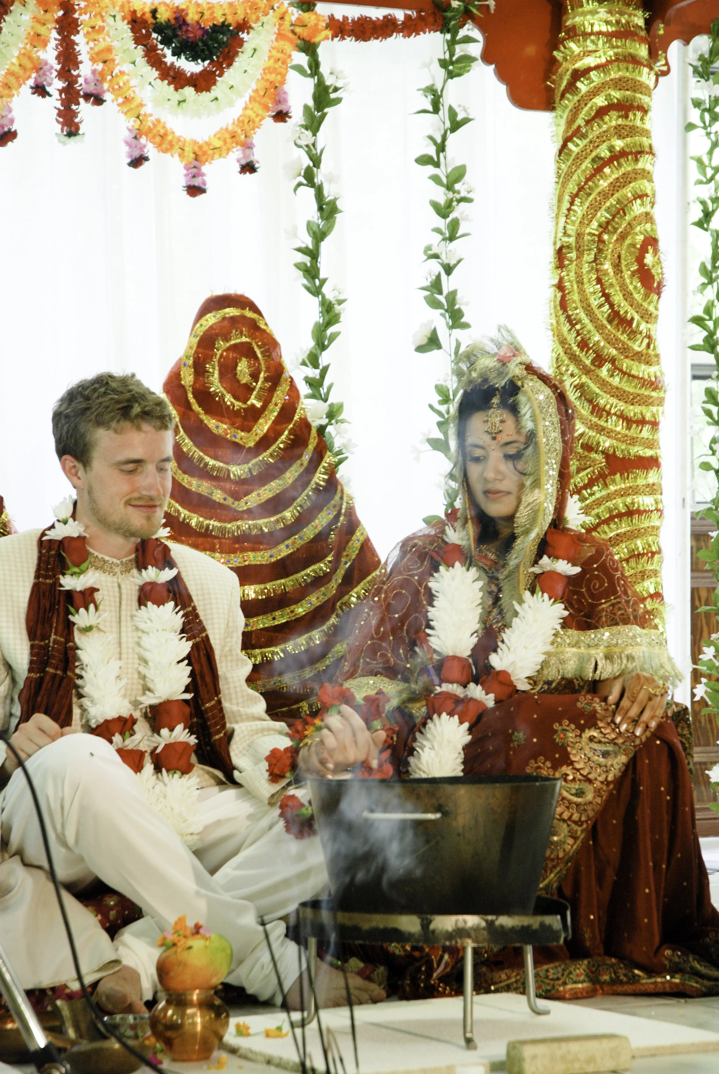 A Hindu wedding ceremony with a south asian bride and caucasian groom sitting on the floor, adorned with flower garlands, surrounded by colourful decorations, including red and gold fabric, flowers, and traditional jewelry in Oakville Ontario.