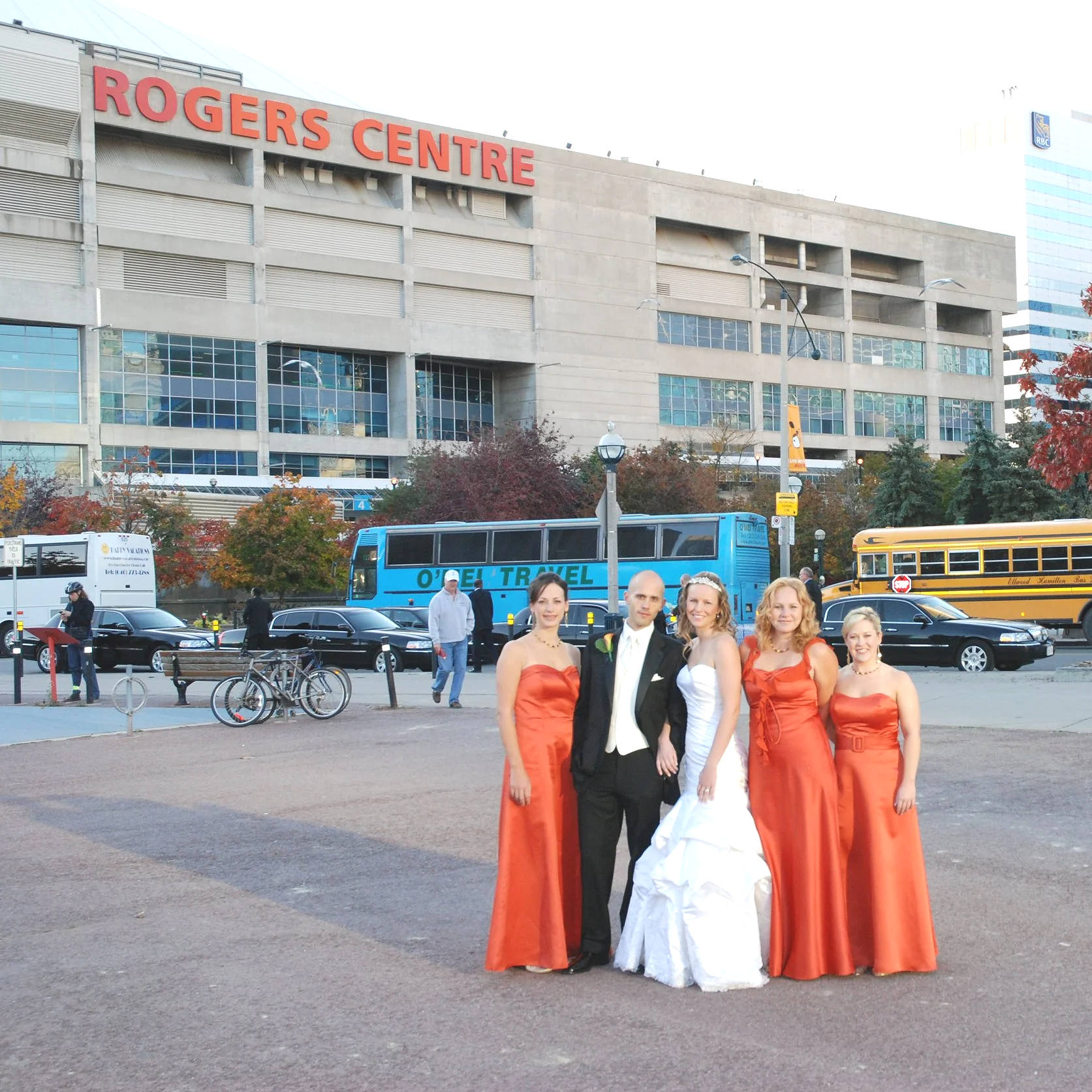 Bride, groom and bridesmaids standing outside the Rogers Centre in Toronto Ontario. Bridesmaids in orange dresses and groom in a black tuxedo,  buses and cars in the background.