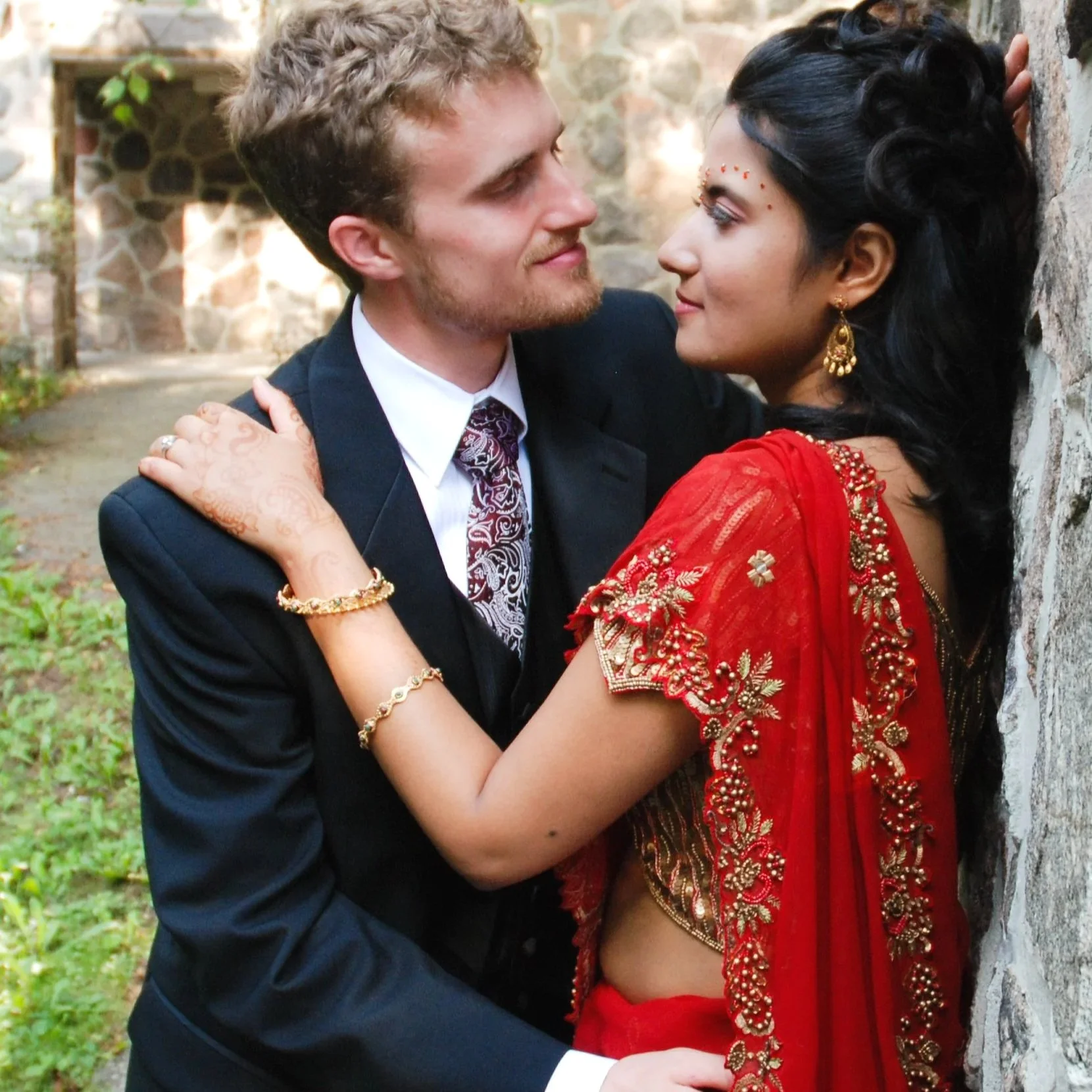 A couple, a white man in a dark suit and an Indian woman in a red traditional Indian saree with gold embroidery, sharing an intimate moment outdoors by a stone wall, gazing into each other's eyes. Kleinburg Ontario.