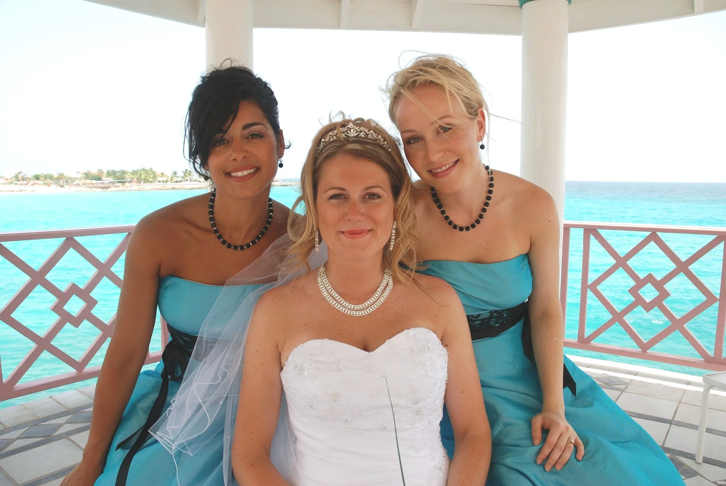 Two bridesmaids in blue dresses leaning in to the bride in a white dress. Tropical ocean in the background. All smiling, sunny day. Destination wedding in Saint Martin.