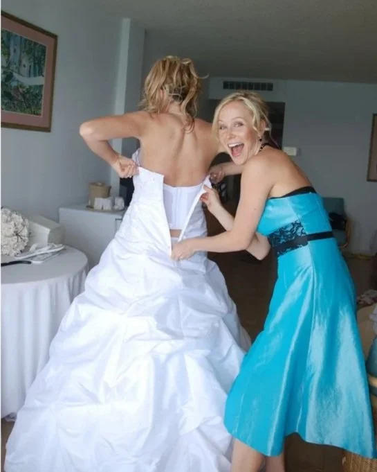 Two women, one in a wedding dress and the other in a blue blue dress, laughing and adjusting the bride's dress in a room in Saint Marten. Destination wedding.