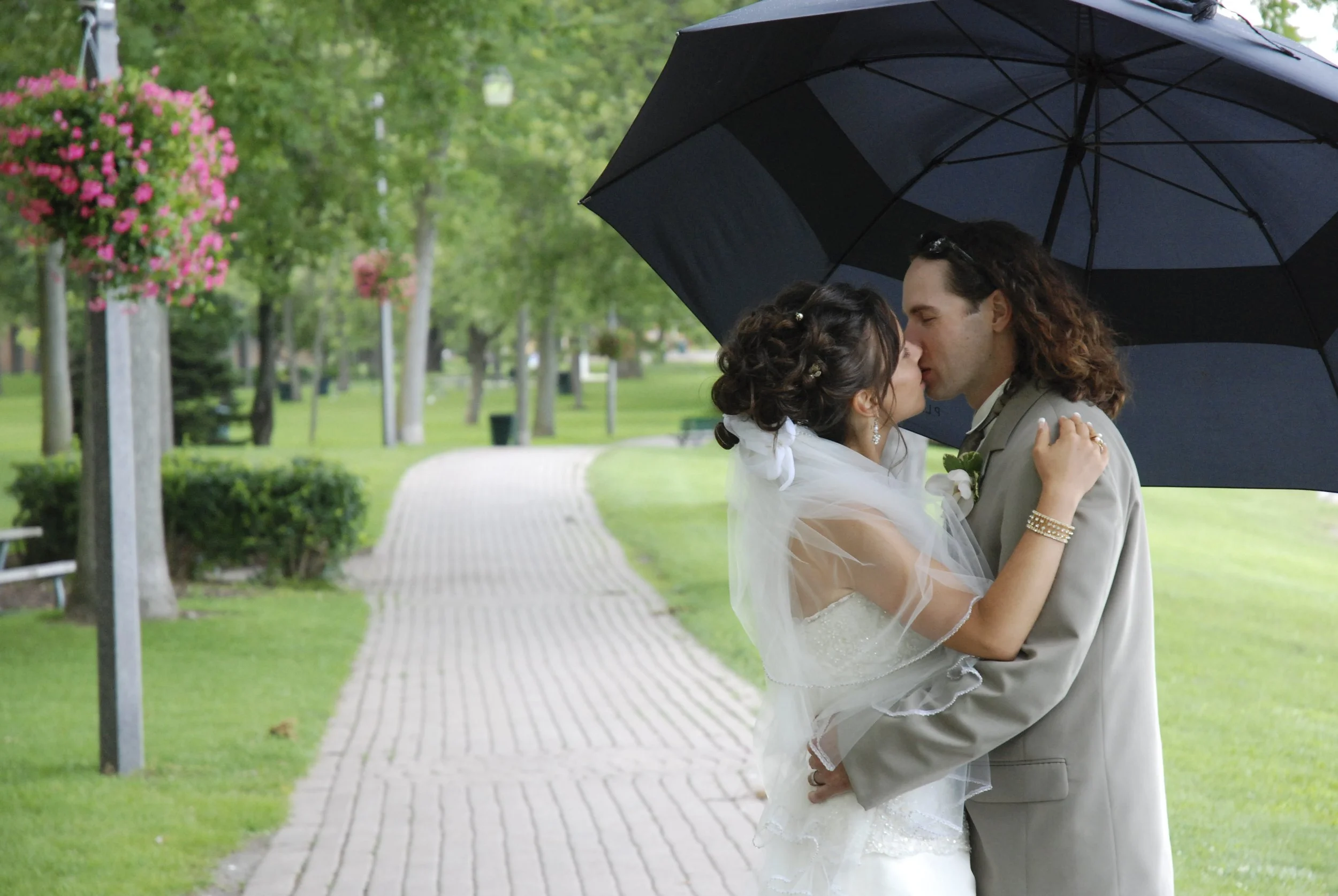 A newlywed couple sharing a kiss under an umbrella in a park with a winding brick path and green trees in Oshawa Ontario.