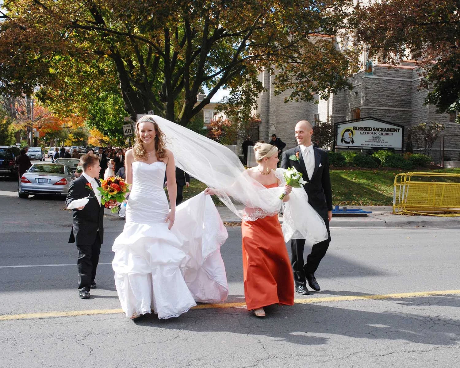 A wedding party crossing the street outdoors in front of a church with a sign that reads Blessed Sacrament Catholic Church in Toronto Ontario, with a bride in a white wedding dress, a groom in a black tuxedo, a bridesmaid in an orange dress,.