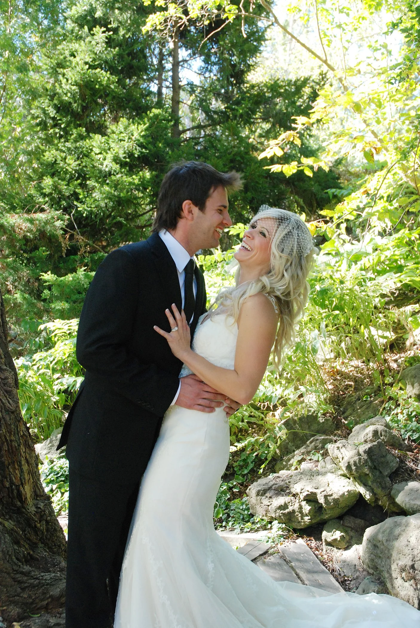 A newlywed couple shares a joyful moment outdoors in a lush green setting. The groom wears a black suit and tie, and the bride wears a white wedding gown with a birdcage veil. They are smiling at each other at Edward Gardens in Toronto Ontario.